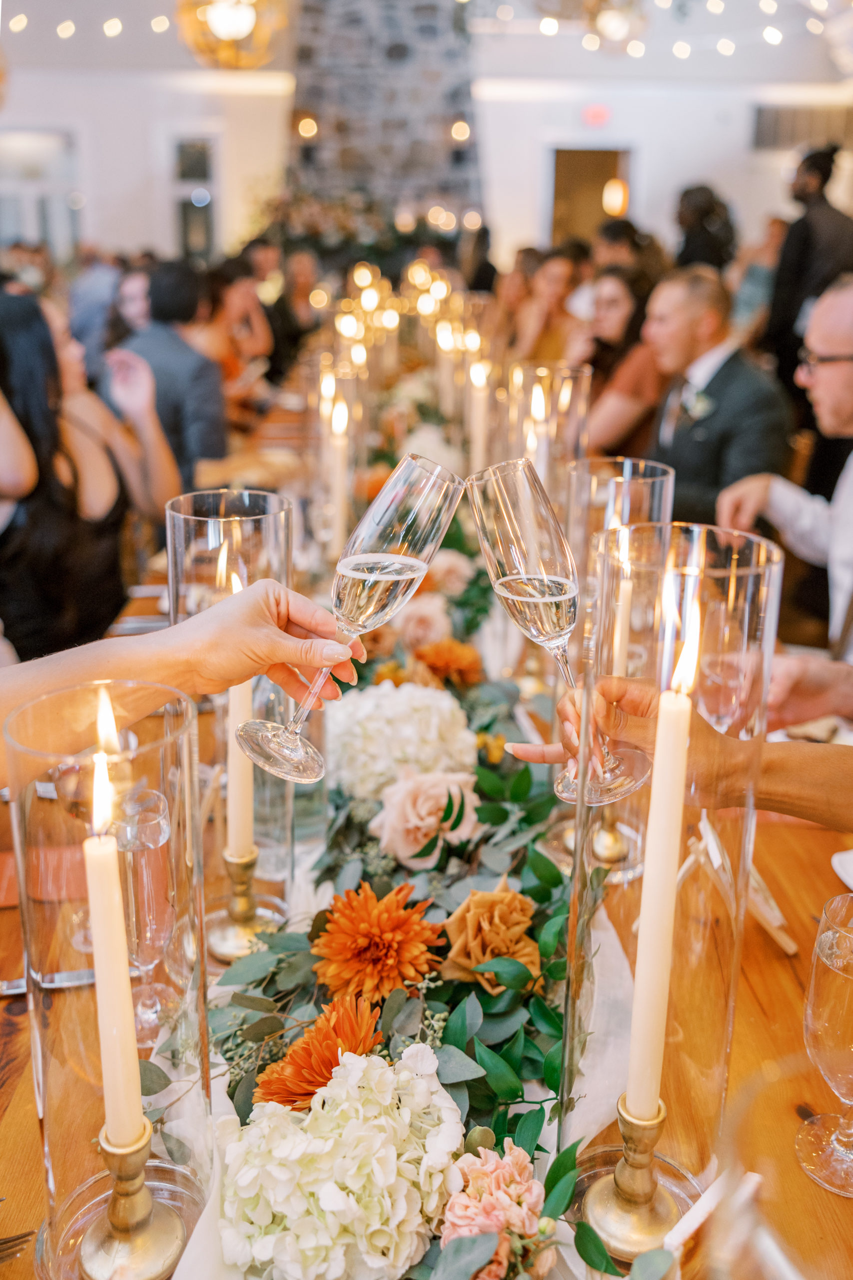 Champagne "cheers" at long wedding reception table with tall candles in vases at this pomme radnor wedding
