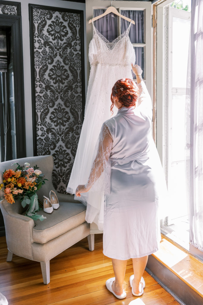 Bride admires wedding dress hanging on the door frame wearing white robe 