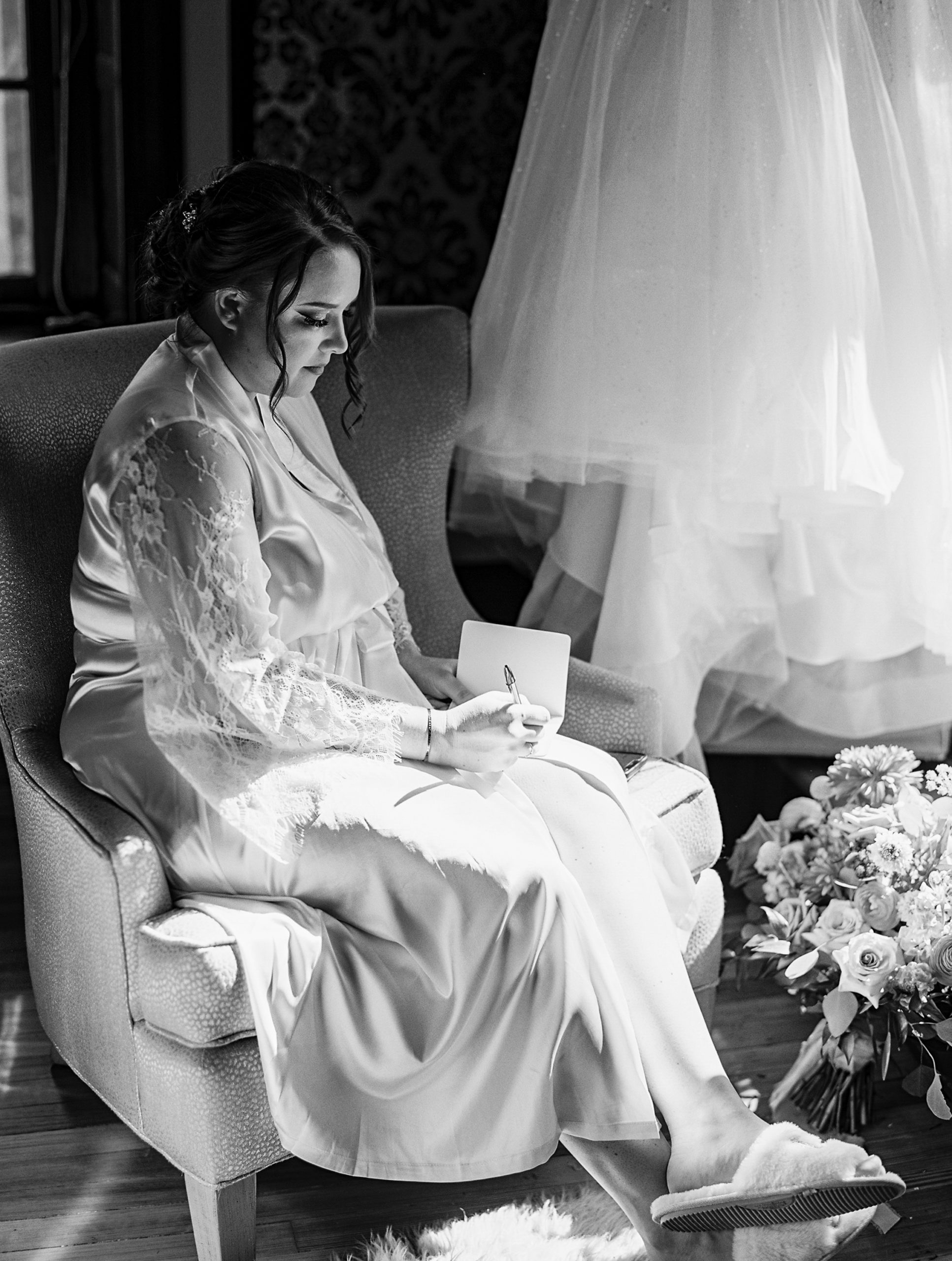 Bride writes letter as she sits on chair with dress hanging up behind her for philadelphia wedding photographer