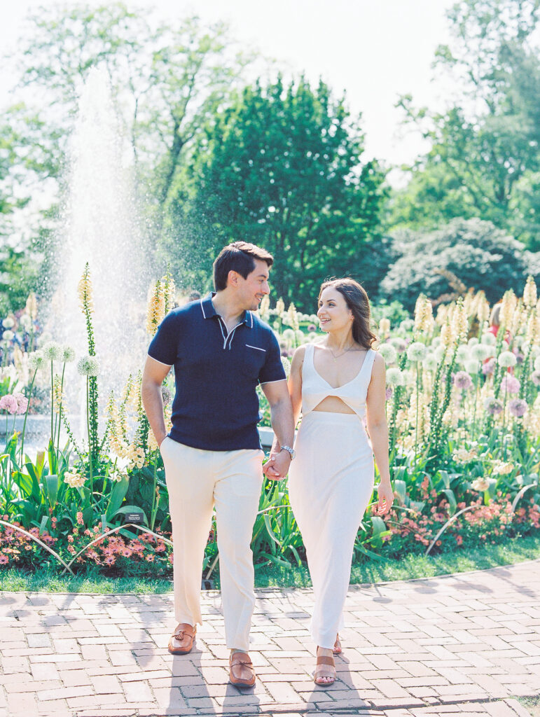 couple walks hand in hand in front of fountain, man wears blue polo shirt and woman wears cream knot front dress