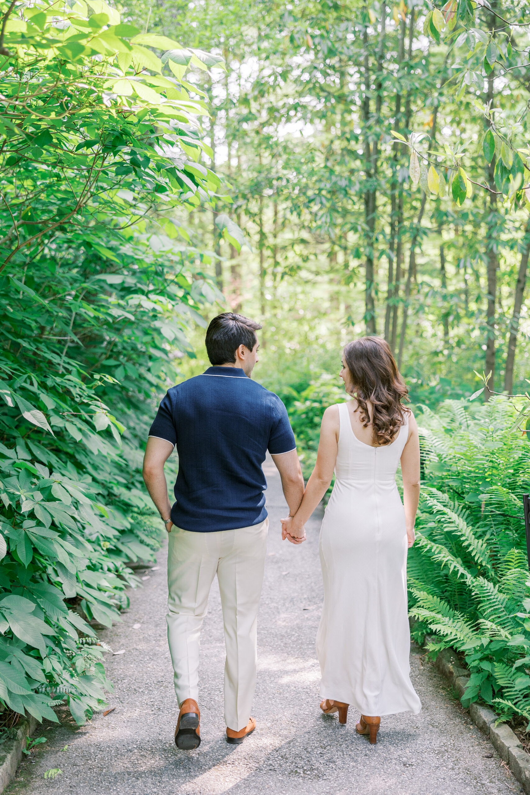 Spring Longwood Gardens Engagement Session shot on film by destination wedding photographer Katie Trauffer - couple walks away hand in hand on sunny tree lined path through dense foliage
