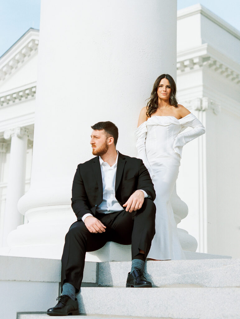 Couple poses on marble steps with large column, man in black suit, woman in white silk engagement dress