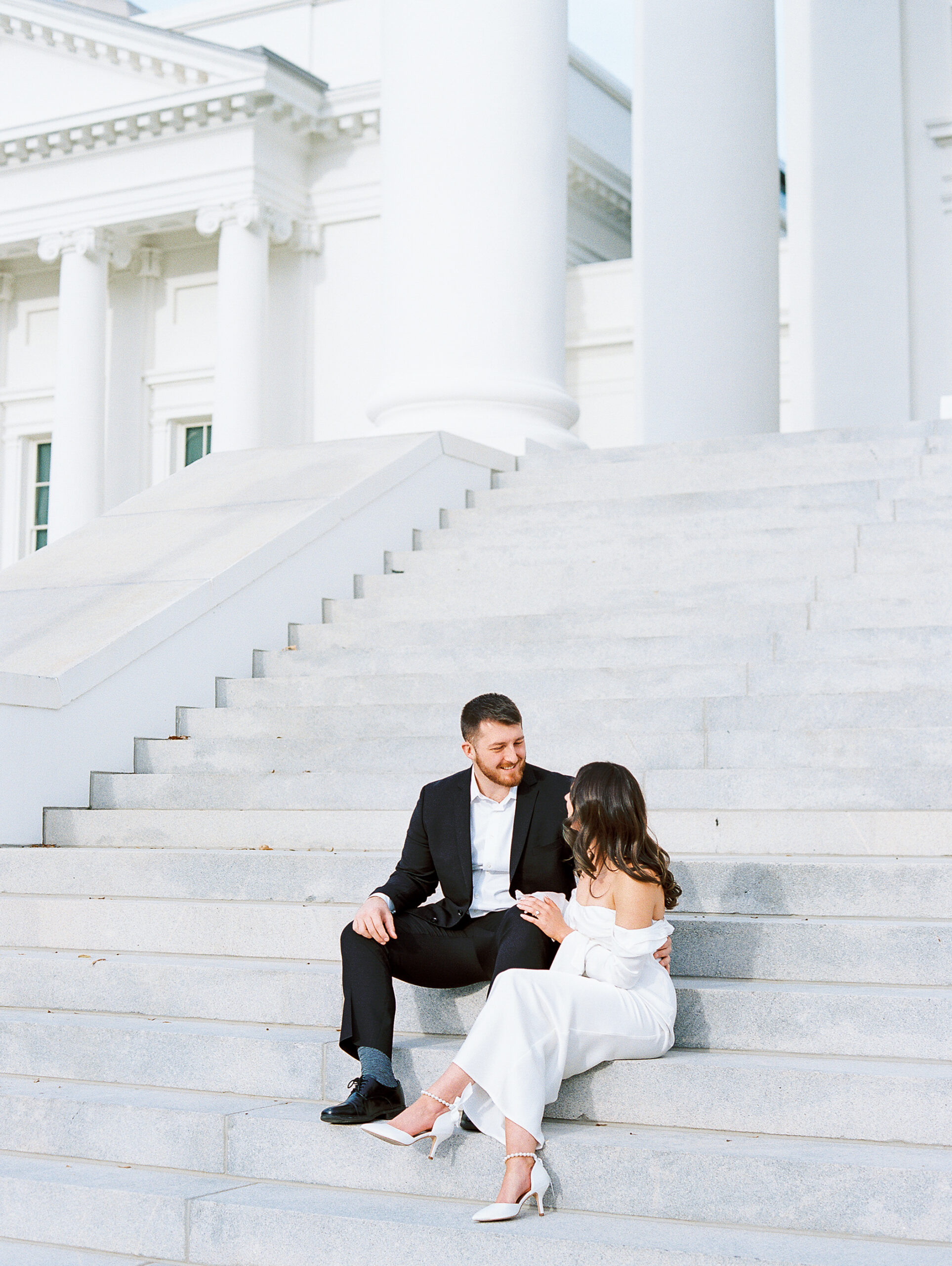 engaged couple sits on marble steps wearing suit and chic white silk dress