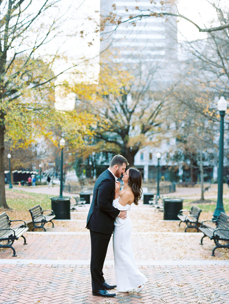 couple embraces on bench lined path with yellow trees and city buildings