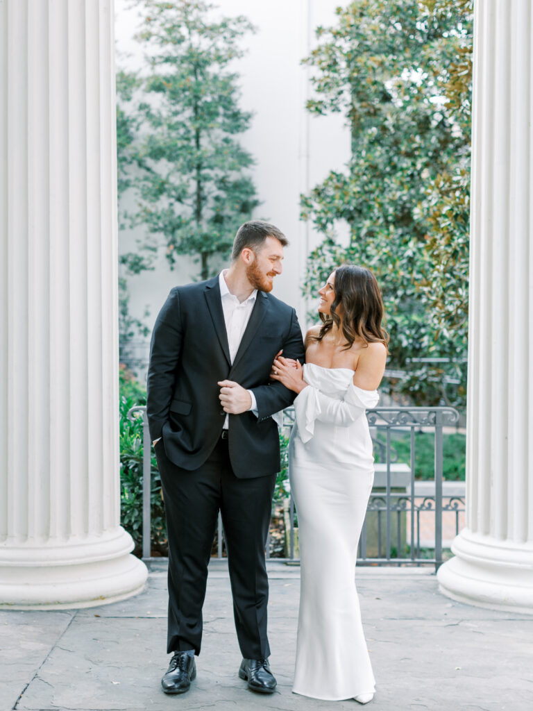 couple walks arm in arm under marble columns with green trees in distance in city setting