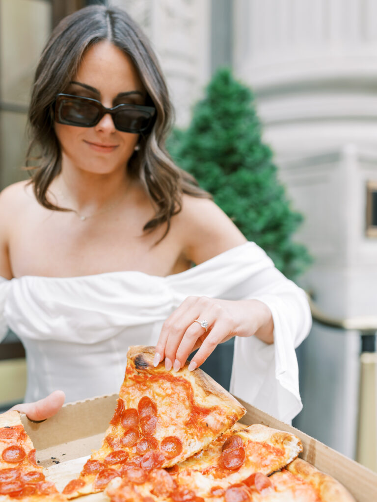 engaged bride wearing white silk dress and sunglasses picks up slice of pizza