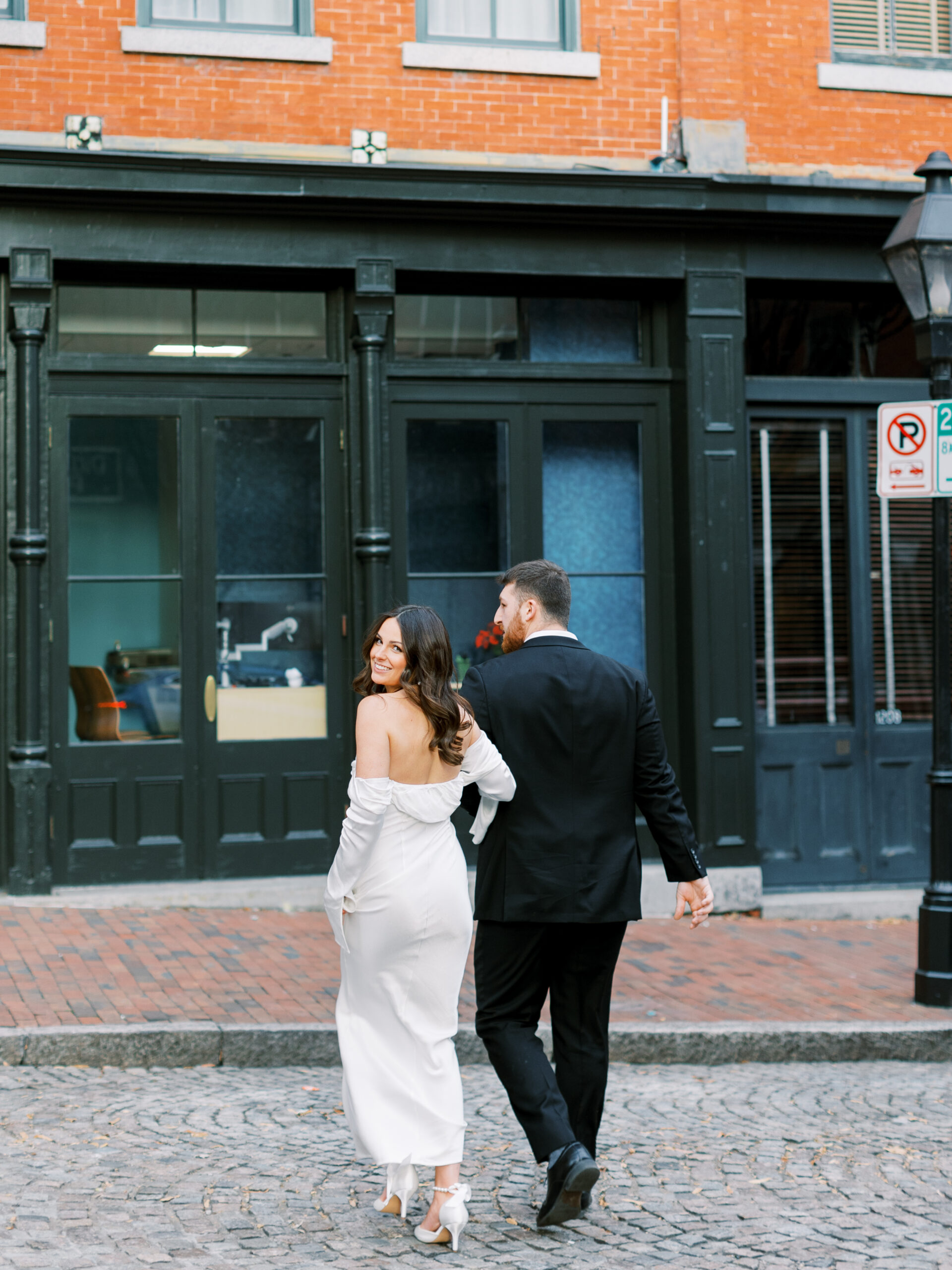 engaged couple in suit and white silk dress cross cobblestone downtown street