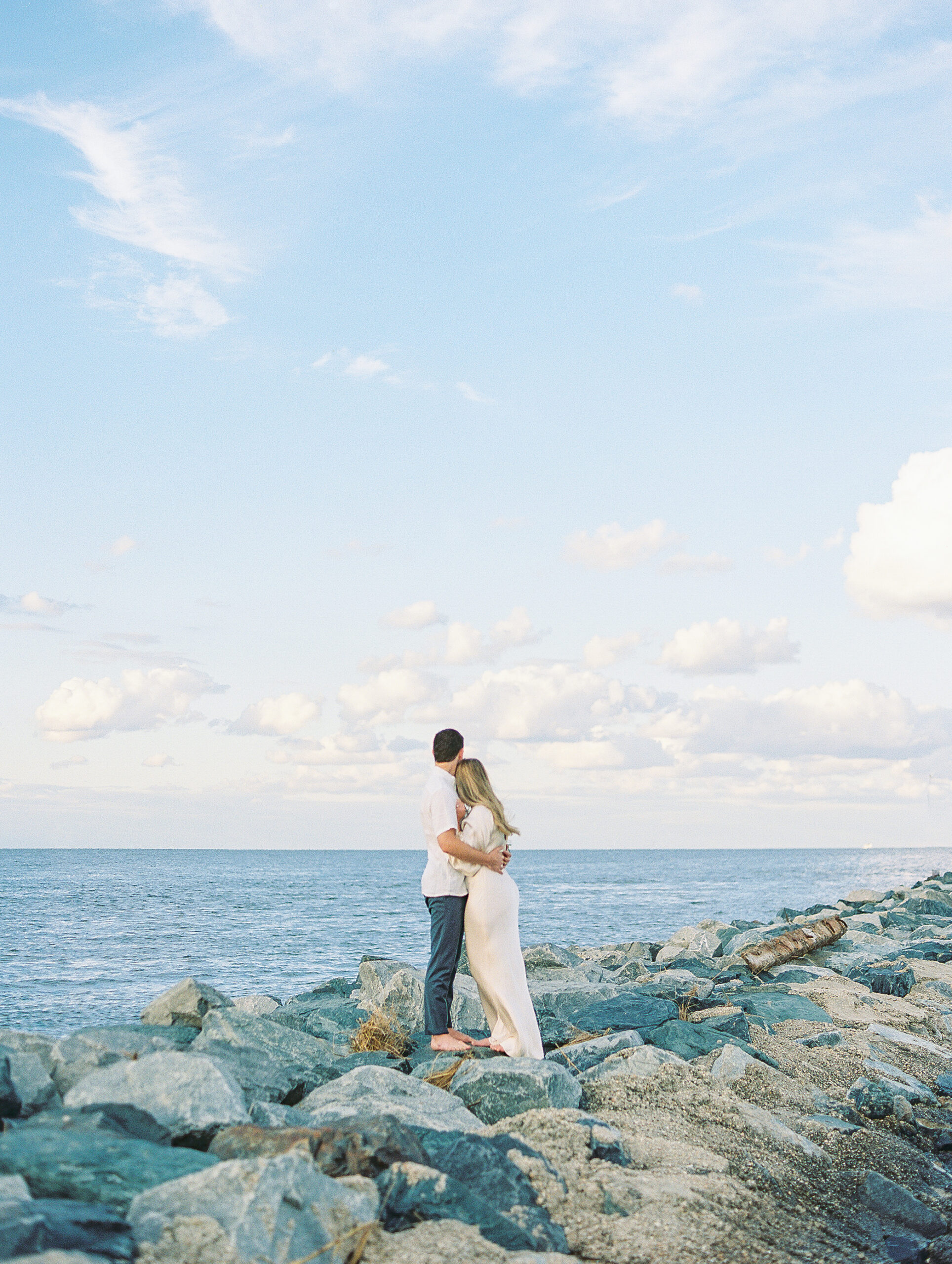 Lewes Beach Engagement Session Couple stands on rocky outcrop in front of blue ocean