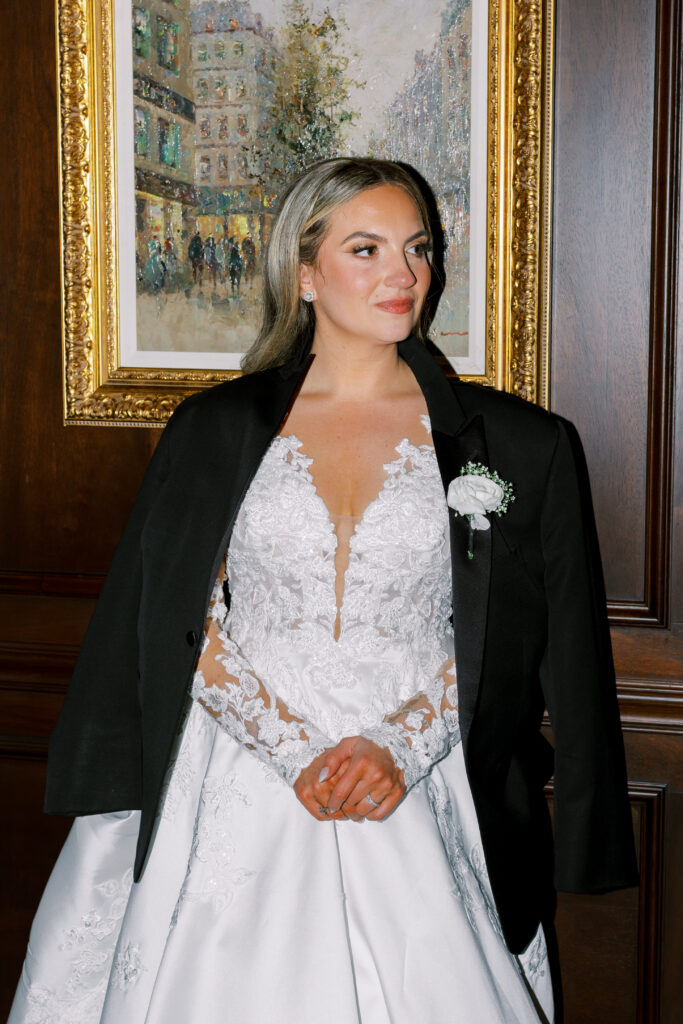 bride stands serenely in dark library wearing groom's jacket