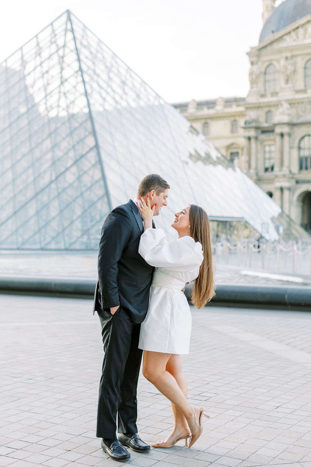 Paris Engagement Photoshoot couple smiles at one another in front of Louvre pyramid